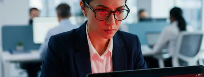 IT-beveiligingsprofessional bekijkt crisisrespons-checklist op laptop, rood alarmlicht op gezicht, collega's op achtergrond.