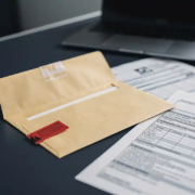 envelop-waarschuwing-documenten-bureau-officieel.jpg Opengevallen manillaenvelop met rode waarschuwingstag op modern bureau, omringd door officiële documenten en gedeeltelijk zichtbare laptop.