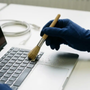 Gehandschoende hand die een zilverkleurig laptoptoetsenbord reinigt met een klein borsteltje, waterdruppels zichtbaar op het oppervlak.