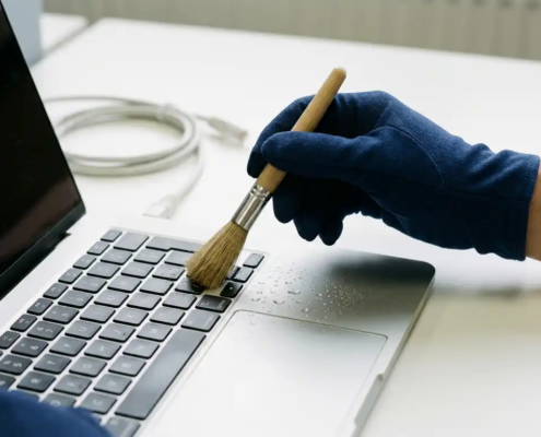 Gehandschoende hand die een zilverkleurig laptoptoetsenbord reinigt met een klein borsteltje, waterdruppels zichtbaar op het oppervlak.
