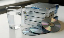 Halfvol glas water naast gestapelde, ongeopende Microsoft-softwaredozen op wit bureau, met cd's die uit een doos vallen.