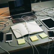 rommelig-bureau-kabels-apparaten-chaos.jpg Rommelig bureau met verstrengelde oplaadkabels, laptop, tablet, drie telefoons, planner en sticky notes in grijs-amberkleurig kantoorlicht.