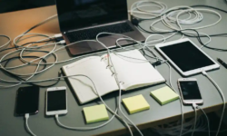 Rommelig bureau met verstrengelde oplaadkabels, laptop, tablet, drie telefoons, planner en sticky notes in grijs-amberkleurig kantoorlicht.