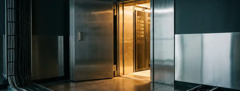 Zware stalen kluisdeur op een kier in een moderne serverruimte, met warm amberkleurig licht dat over betonnen vloer valt.