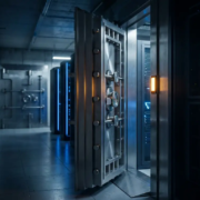 kluis-serverruimte-stalen-deur-beveiliging.jpg Zware stalen kluisdeur op een kier in een moderne serverruimte met blauw omgevingslicht en een tweede versterkte deur op de achtergrond.