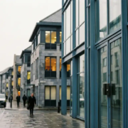toeleveringsketen-beveiliging-kantoorgebouwen-regelgeving.jpg Rij verbonden kantoorgebouwen in staal- en grijstinten met beveiligingsslot op voordeur, symboliseert toeleveringsketenbeheer en regelgeving.
