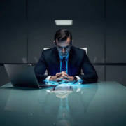 zakelijk-directeur-vergaderzaal-laptop-druk.jpg Zakenman in donker pak aan glazen vergadertafel, verlicht door koud blauw laptopscherm in donkere boardroom.