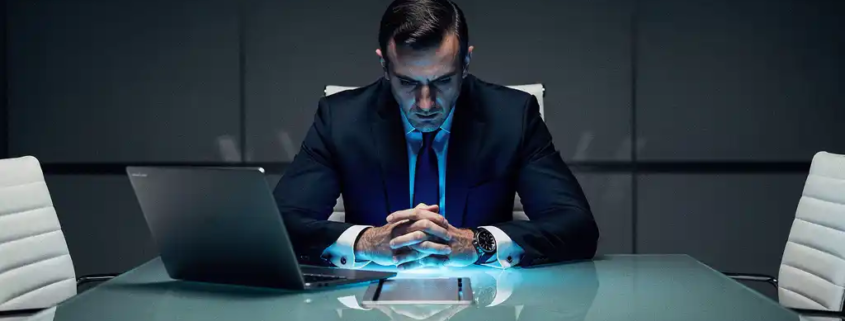 Zakenman in donker pak aan glazen vergadertafel, verlicht door koud blauw laptopscherm in donkere boardroom.