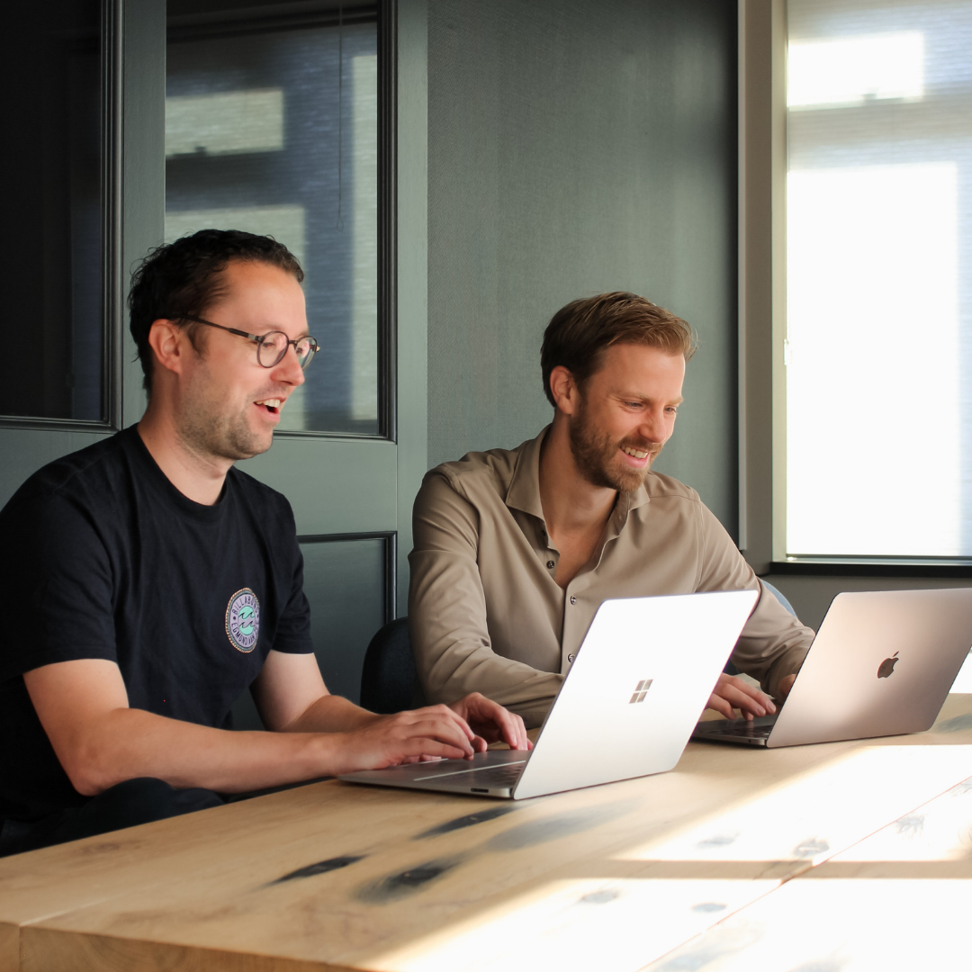 Copilot said: Twee medewerkers werken samen achter een laptop in een vergaderruimte en bespreken informatie op het scherm.