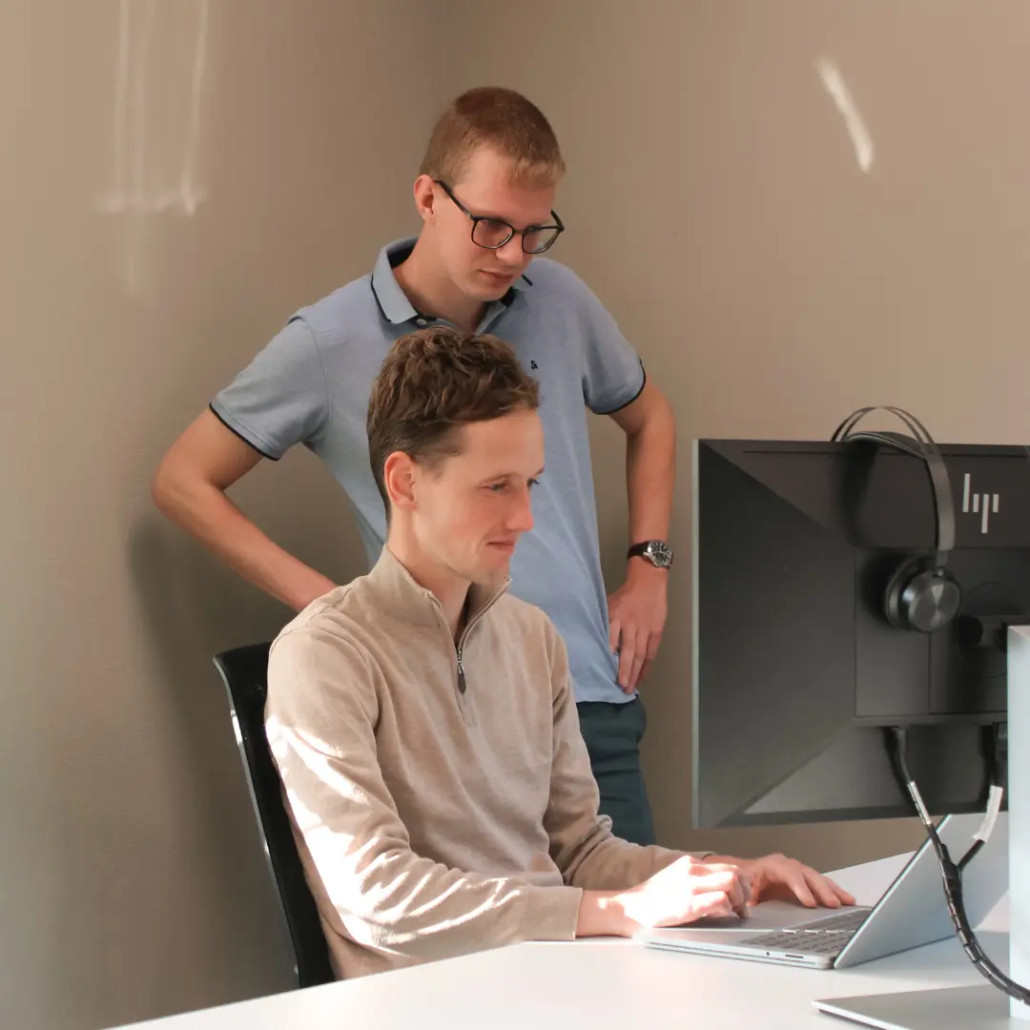 Microsoft Power Platform Twee medewerkers van Puur ICT kijken samen naar een computerscherm, één zittend en één staand achter het bureau.