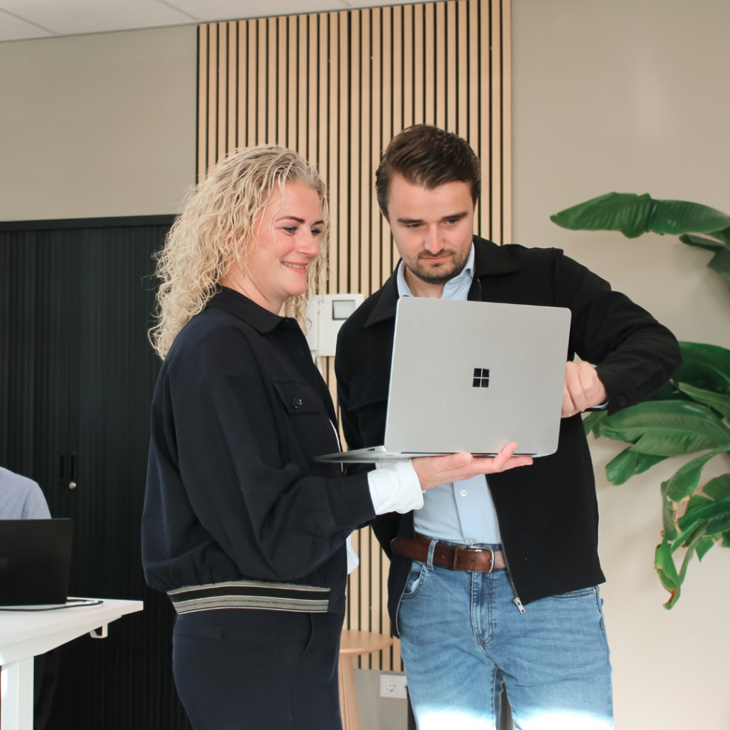 Twee collega's staan samen op kantoor en kijken aandachtig naar een laptopscherm. Ze overleggen terwijl ze door informatie op het scherm gaan in een moderne werkomgeving.