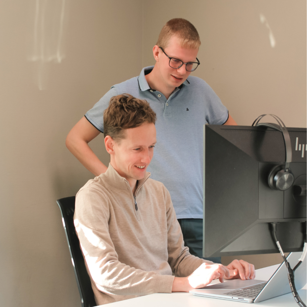 Een collega staat achter een zittende collega en kijkt mee naar het scherm van diens laptop. Samen bespreken ze informatie op het scherm in een kantooromgeving.