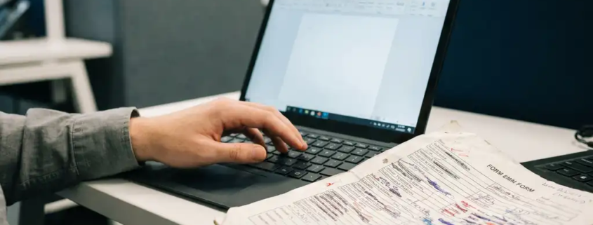 Gefrustreerde kantoormedewerker boven doorgestreept papieren formulier met inktvlekken, laptop op achtergrond als digitaal alternatief.